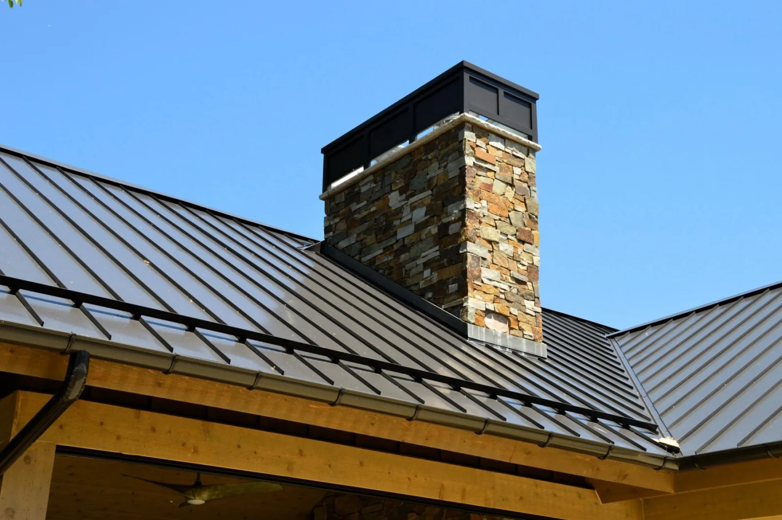 Standing-seam metal roof with a natural stone chimney and timber soffit against a clear sky — Iconic Construction project